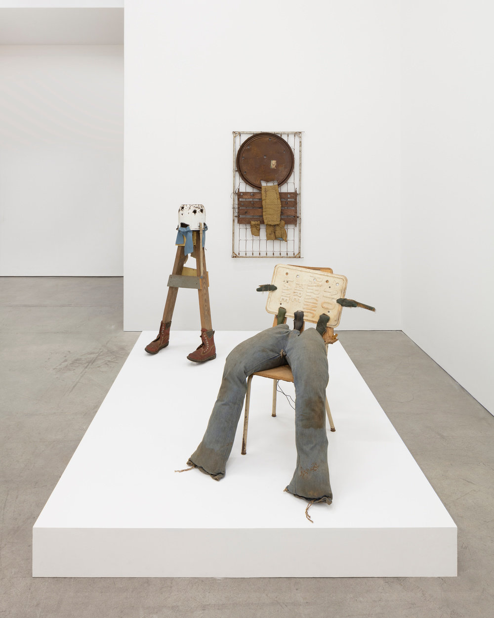 An installation view of three sculptures of found-object construction by Hawkins Bolden. Two sculptures appear as anthropomorphic figures on a white platform base. The first in the foreground sits on a wood seat school chair. A faded "No Parking" sign makes the torso of the figure with a strip of tire protruding on the left and right edges. Muddied pants extend down from the chair and are stuffed to give them form. On the back end of the platform the sculpture appears standing on two 4" x 4" wood planks fitted with brown leather work boots. A rusted pot sits upside down on the wood planks and is cut with holes that appear as a face. Blue leather swatches hang from the mouth of the pot and other areas around the rim of the pot. The third sculpture appears mounted on the back gallery wall. 