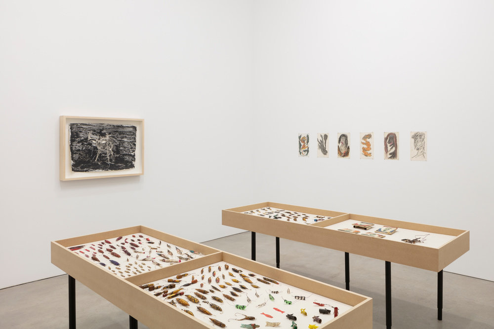 An installation view of work by Thornton Dial. Fishing lures are displayed on two tables and drawings are mounted on the right back wall. "Deer" by Ronald Lockett is mounted on the back left wall.