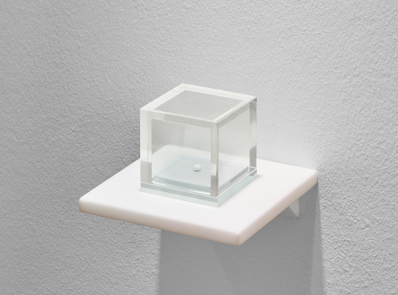 A small scale sculpture of a glass cube with a water droplet inside that sits on a white shelf by Lars Fisk. 