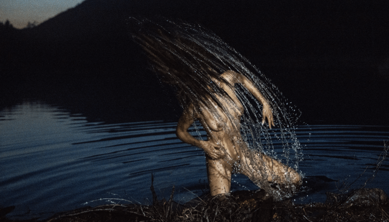A color photograph by Ryan McGinley of a nude woman in nature, creating a spray of water as she throws her head back. 