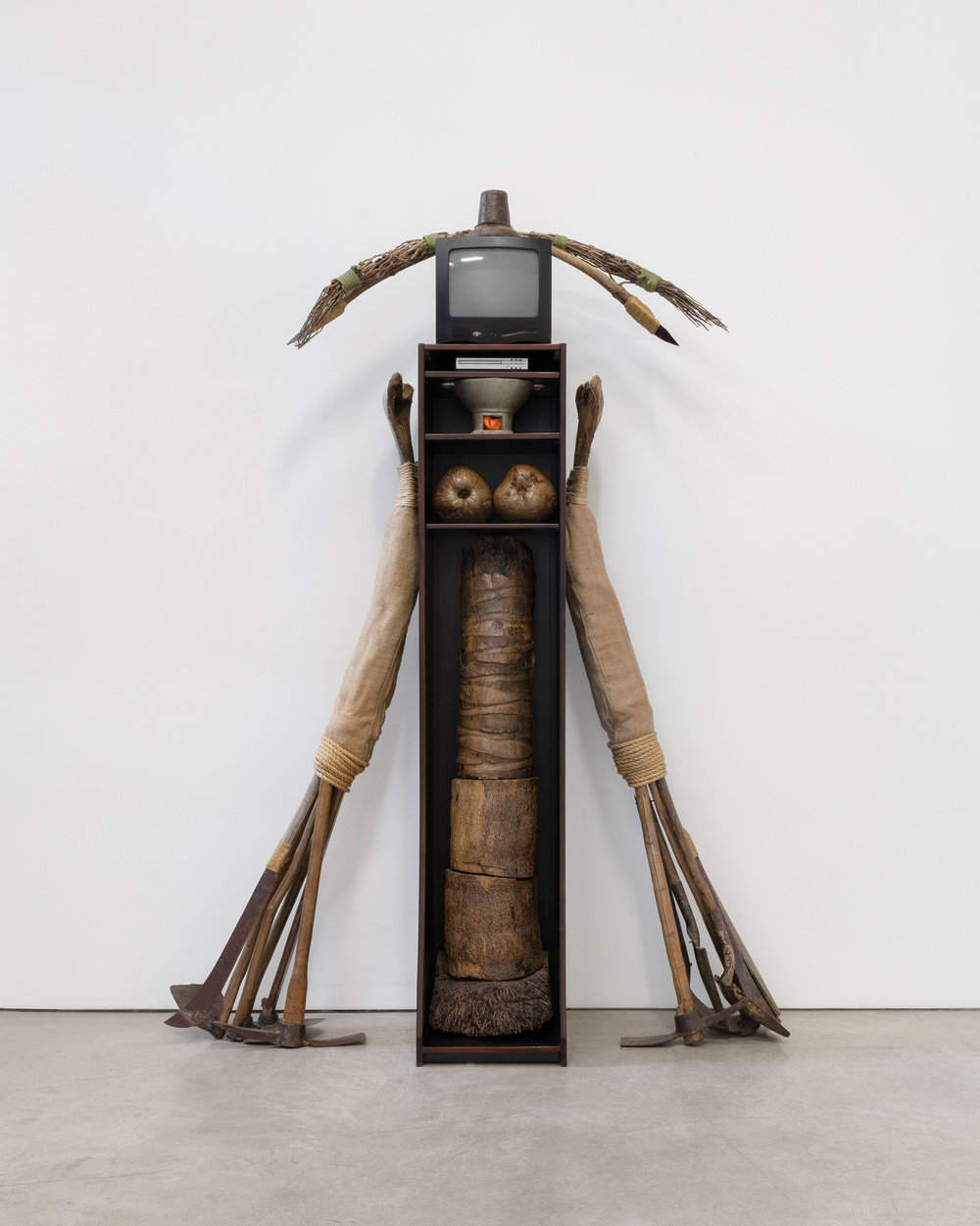 A large, mixed media sculpture by Daniel Lind-Ramos made of a pick, shovel, machetes, steel, palm tree trunks, dried coconuts, TV monitor, palm tree branches, DVD player, wood panel and fabric.