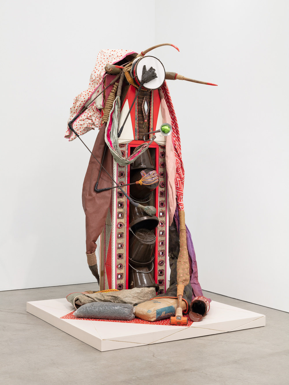 A mixed media sculpture by Daniel Lind-Ramos made of mirrors, concrete blocks, cement bag, sledge hammer, construction stones bag, paint bucket, wood panels, palm tree trunk, burlap, leather, ropes, sequin, awning, plastic ropes, fabric, trumpet, pins, duct tape, maracas, sneaker, tambourine, working gloves, basketballs, boxing gloves and acrylic.